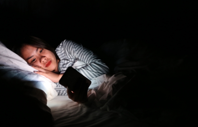 Blonde female man looking at bright phone in bed 