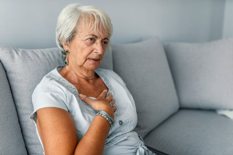 A woman is sitting on her couch feeling something out of breath and clutching at her chest.